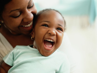 Mãe, de pele negra, sorrindo e segurando sua bebê, também de pele negra, que está sorridente e veste uma camiseta azul clara