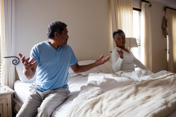 casal de idosos sentados em lados opostos da cama durante discussão, o homem está com os braços abertos e a mulher apontando o dedo