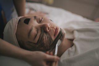 Jovem mulher deitada em cama com máscara de carbono no rosto sendo aplicado com espátula por esteticista