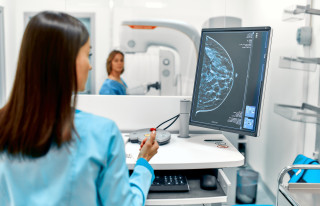 Médica analisando um resultado de mamografia ao mesmo tempo em que a paciente realiza o exame