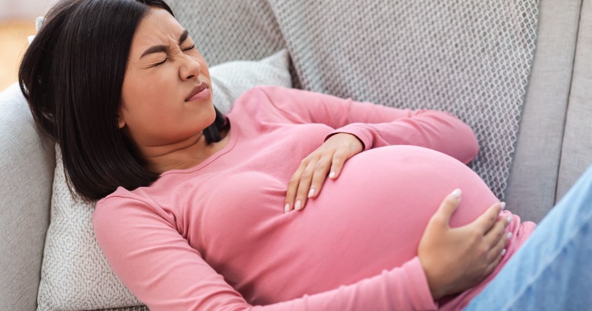 mulher grávida deitada em sofá segurando a barriga com as mãos e sentindo dor