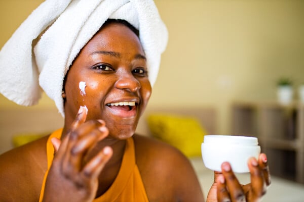 mulher negra aplicando creme no rosto e usando uma toalha branca no cabelo