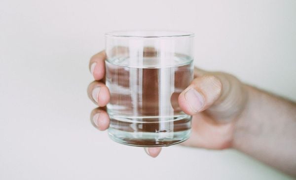 Close up da mão de uma pessoa segurando um copo com agua em frente a uma parede branca