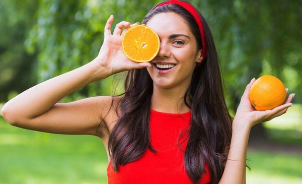 Mulher sorrindo segurando duas laranjas: com a mão direita coloca uma no olho e com a mão esquerda exibe a laranja