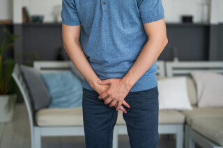 Homem veste calça jeans e camiseta azul e cobre a região genital com as mãos. A dor é um dos sintomas de herpes genital