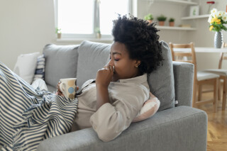 Jovem mulher, de pele negra e cabelos crespos pretos, sentada no sofá assoando o nariz e segurando uma caneca com chá