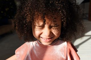 criança negra de cabelo cachecado com os olhos fechados e sorrindo, usando uma blusa rosa