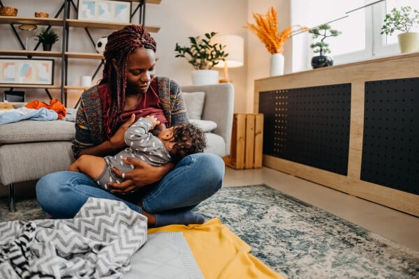 Mulher negra amamentando um bebê enquanto está sentada no chão de uma sala
