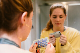 Mulher usando blusa amarela na frente do espelho segurando uma cartela de anticoncepcional