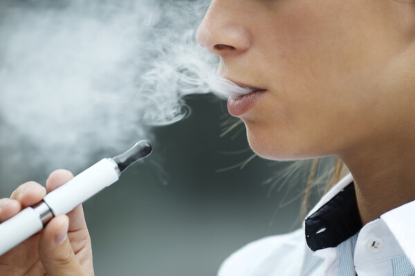 Imagem aproximada de mulher soltando fumaça pela boca enquanto segura com uma das mãos um cigarro eletrônico