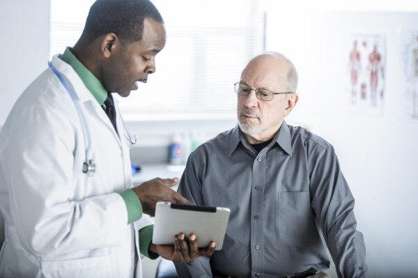 Homem branco com barba sendo atendido em consultório por médico negro mostrando tablet