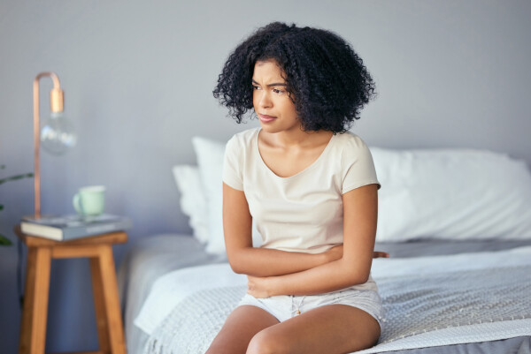 Mulher negra, com roupas claras e cabelos cacheados, sentada na ponta da cama com os braços envolta do abdômen, sentindo cólicas, sintomas de endometriose