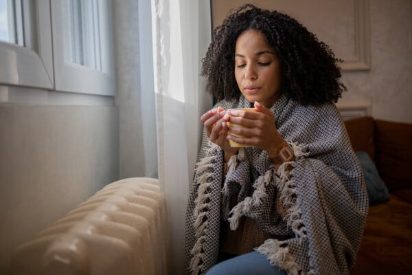 mulher de olhos fechados sentada no sofá de sala, na frente de uma janela, segurando uma xícara de chá com as mãos