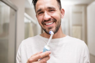 Homem segurando uma escova elétrica e sorrindo na frente do espelho. Ele tem cabelo preto, barba e está vestindo uma camiseta branca. O fundo da imagem é o banheiro.