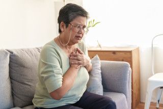 Mulher idosa sentada em sofá de óculos com as duas mãos no peito sentindo dor