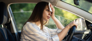Mulher caucasiana dirigindo um carro com a mão esquerda na testa indicando calor excessivo