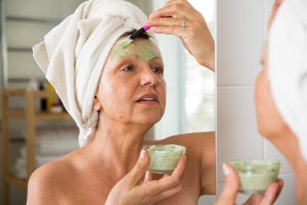 mulher na frente do espelho aplicando uma máscara facial verde com uma espátula