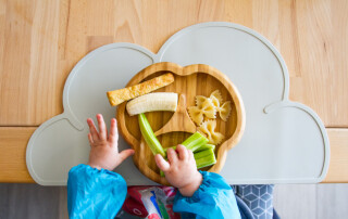 Close visto de cima das mãos de um bebê sentado em um cadeirão mexendo nos alimentos que estão em um prato
