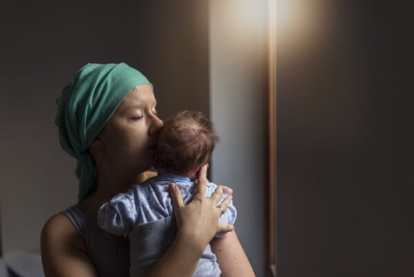 Paciente com câncer usa lenço verde na cabeça enquanto segura seu bebê no colo e o beija no rosto
