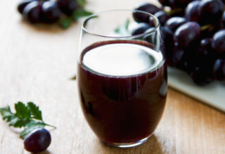 Copo de vidro com suco de uva vermelha e uvas ao lado. 