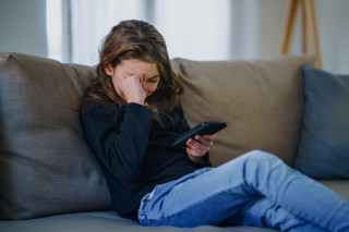 Criança sentada no sofá com celular na mão e chorando
