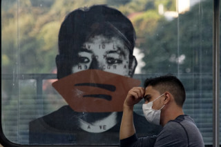 Homem usando máscara protetora branca, sentado no metrô de São Paulo. Ao fundo, uma ilustração de um rapaz usando máscara protetora.