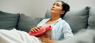 Mulher sentada em um sofá segura uma bolsa de água quente na região do abdômen e faz expressão de dor
