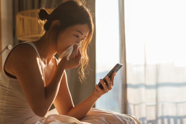 Mulher asiática, sentada na cama com lençóis brancos, assoando o nariz com um lenço e mexendo no celular com a outra mão