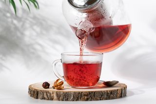 Chá de hibisco sendo despejado de uma jarra de vidro para uma xícara de vidro; ao lado uma colher e hibiscos