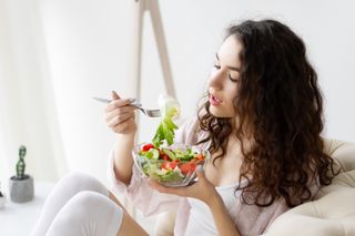 Mulher comendo uma salada de alface e tomate enquanto está sentada no sofá