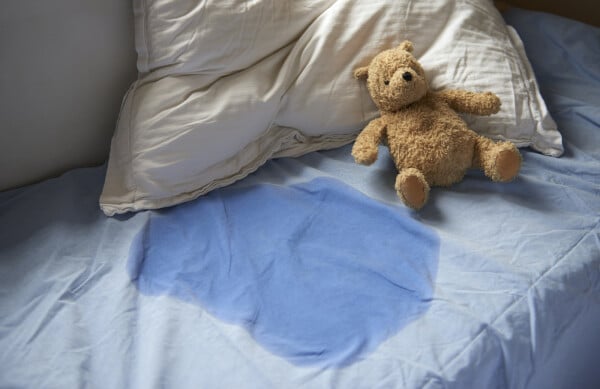 Cama com lençol azul e travesseiro branco com mancha de xixi no meio e um ursinho de pelúcia ao lado