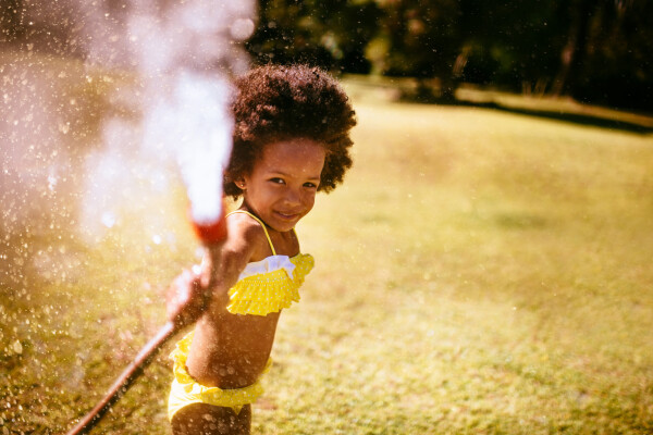Criança de pele negra e cabelos crespos castanhos veste biquíni amarelo e joga água de uma mangueira em um jardim