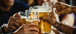 Foto aproximada de cinco mãos segurando um copo de cerveja, brindando.