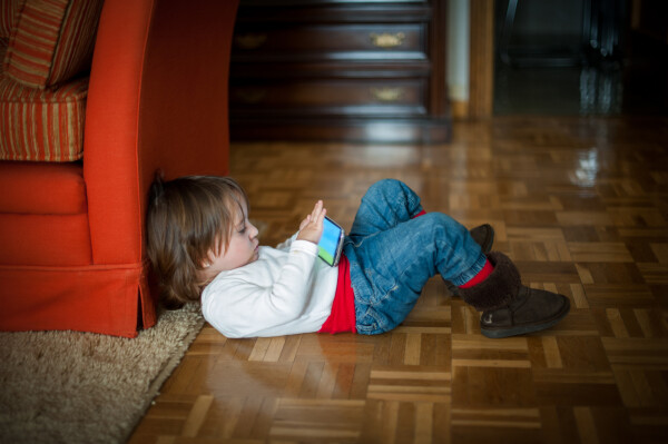 Criança deitada no chão, com a cabeça encostada em um sofá, enquanto mexe no celular