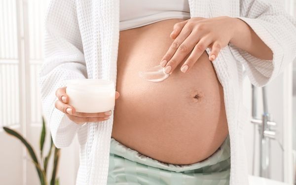 Mulher grávida passando um creme na barriga enquanto segura o pote com uma mão