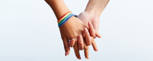 Foto aproximada de duas mãos juntas. Uma delas usa pulseiras com as cores da bandeira LGBTQIAP+.