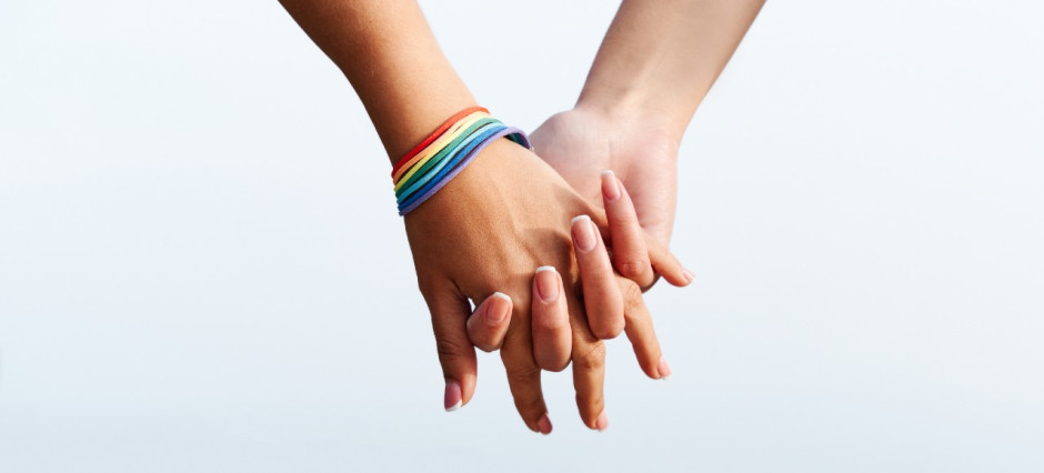 Foto aproximada de duas mãos juntas. Uma delas usa pulseiras com as cores da bandeira LGBTQIAP+.