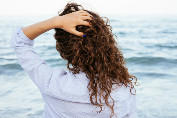 Imagem de uma mulher de costas para a câmera, mas de frente para o mar, e com a mão no cabelo ondulado