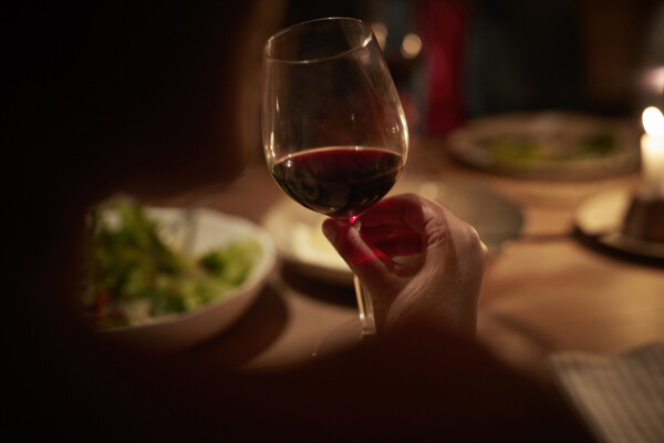 Foto aproximada de mão feminina, de pele branca, segurando uma taça de vinho tinto; ao fundo, pratos com salada