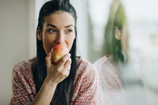 Mulher ao lado de janela comendo uma maçã