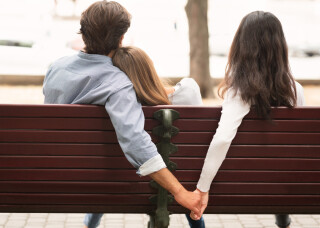Homem sentado ao lado de sua parceira abraçada a ele e segurando a mão de outra mulher ao lado do casal