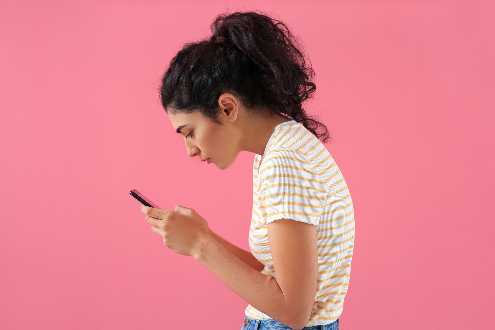 Jovem mulher de cabelos pretos encaracolados mexe no celular enquanto apresenta coluna curvada