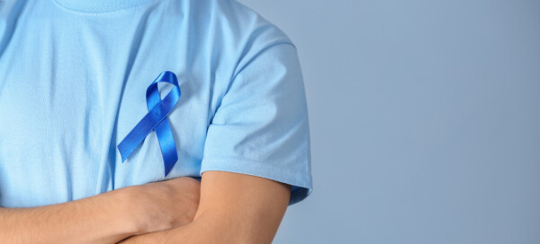Homem de camiseta azul e braços cruzados com a fita símbolo do novembro azul pregada no peito