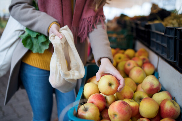 Mulher comprando maças em uma feira, segurando uma maçã na mão e um saco branco na outra mão