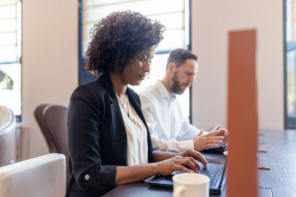 Mulher, vestindo blazer preto e camiseta branca, sentada em uma cadeira e digitando em seu notebook, ao lado de um homem de barba e camisa branca