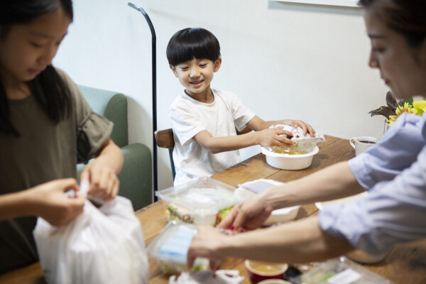 Família japonesa, composta por mãe, filha e filho pequenos, abrindo embrulhos de comida em cima de uma mesa de madeira