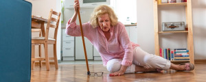 Mulher idosa loira vestindo camisa rosa e calça branca no chão apoiada em sua bengala. Seu óculos está no chão.