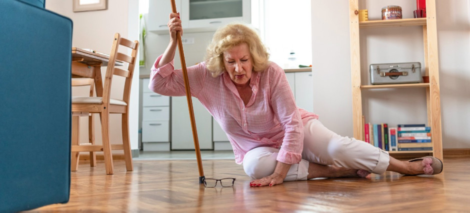 Mulher idosa loira vestindo camisa rosa e calça branca no chão apoiada em sua bengala. Seu óculos está no chão.