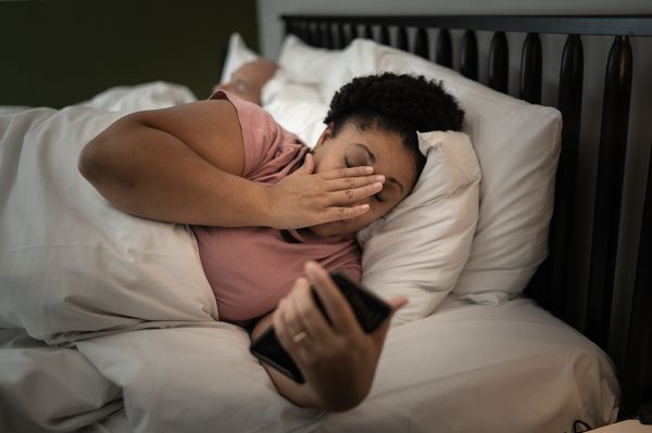 Mulher negra deitada na cama com um celular na mão. Sua expressão é de sonolência e estresse