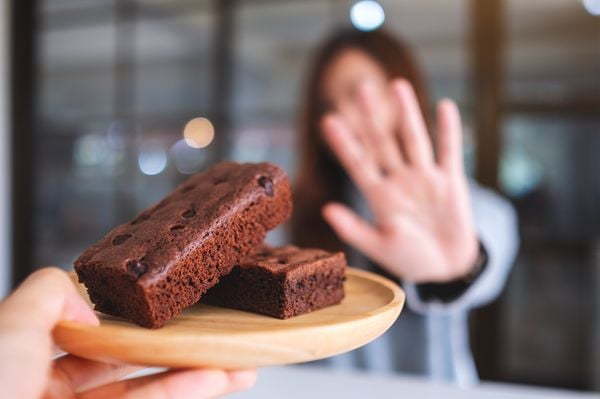 Mulher desfocada ao fundo recusando um bolo de chocolate que está em primeiro plano